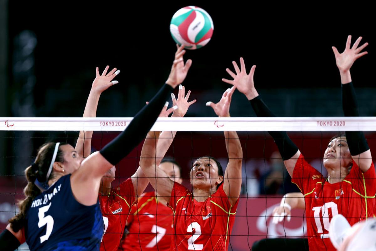 A female athlete tips the ball over the net, while three players try to block it